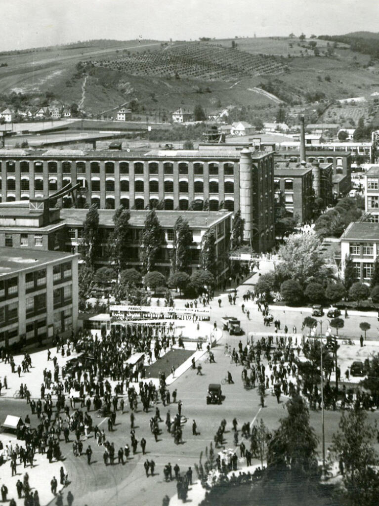 Zahlreiche Menschen strömen in das Gelände der Bat´a Fabrik mit mehreren unterschiedlichen Gebäuden. Postkarte aus den 1930ern.